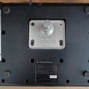 Fotografie zachycuje spodní stranu černého elektronického zařízení. Uprostřed je kovový mechanismus s nápisem Roland. Ve spodní části se nachází štít…