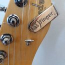 Detail hlavy elektrické kytary z přírodního dřeva. Na dřevě je viditelné částečné logo Fender. Na strunách je upevněn kovový stahovací kousek s nápis…