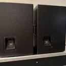 Dva černé reproduktorové boxy s texturovaným povrchem umístěné na transportním case. Na zadní straně každého boxu je v prohlubni umístěn konektor a š…