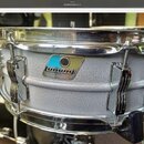 Stříbrný snare drum s texturovaným povrchem a chromovanými obručemi. Na korpusu je viditelný štítek s logem ludwig a nečitelným nápisem pod ním. Nást…