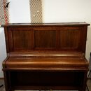 Hnědé dřevěné piano s vertikálním provedením. Povrch vykazuje známky opotřebení a drobné škrábance, zejména v horní části nad klávesnicí. Na nástroji…