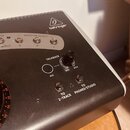 Detailní pohled na horní panel černého elektronického zařízení se stříbrným okrajem. Na povrchu je logo a nápis behringer, sekce MONITOR SELECT s tla…
