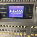 Digitální mixovací pult s šedým povrchem. Uprostřed je modrý displej s bílými nápisy „02R“, „DIGITAL RECORDING CONSOLE“, „Version 2“ a „Copyright (C)…
