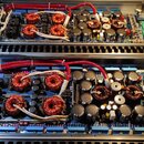 Fotografie zachycuje vnitřek dvou otevřených elektronických zařízení, pravděpodobně zesilovačů. Jsou vidět velké měděné tlumivky, množství černých el…