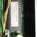 Detailní pohled na zelenou základní desku s elektronickými součástkami. Uprostřed je umístěn černý integrovaný obvod s bílým štítkem, na kterém je te…