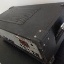 Černý přenosný kufřík s elektronickým zařízením uvnitř, vybavený kovovými rohy a madlem. Horní strana má ventilační mřížku s otvory. Zadní panel obsa…