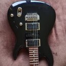 Černá elektrická kytara s lesklým povrchem a hnědým hmatníkem. Na mostku je viditelný nápis Tune. Nástroj má dva humbuckery s kovovými rámečky a jede…
