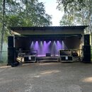 Fotografie zachycuje venkovní zastřešené pódium s osvětlením a zvukovou technikou. Po stranách jsou umístěny černé reproduktorové sestavy a v popředí…
