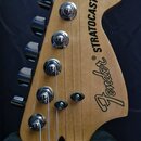 Fotografie zachycuje hlavu elektrické kytary z přírodního světlého dřeva. Na povrchu je černý nápis Fender® STRATOCASTER®. Nástroj má šest chromovaný…