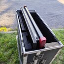 Černý transportní case s hliníkovými hranami a úchyty, v němž je umístěn dlouhý černý tubus s červenými koncovými zátkami. Vnitřek case je vyložen če…