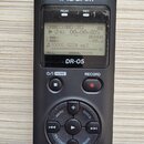 Černý digitální záznamový systém s dvěma stříbrnými mikrofony v horní části označenými písmeny L a R. Na přístroji je bílý nápis TASCAM, modelové ozn…