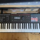 Černý digitální klavír s bílými a černými klávesami. Na pravé straně je nápis Roland JUNO-DS a modrá sekce s označením PHRASE PAD. V horní části jsou…