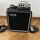 Černý kytarový kombozesilovač s bílým rámečkem a nápisem VOX na přední mřížce. Na horní části přístroje stojí malý zesilovač s kovovou rukojetí, nápi…