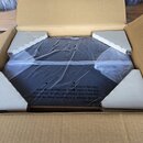 V otevřeném kartonovém boxu se nachází černý předmět s hranatým tvarem, který je zabalen v průhledné plastové fólii. Na fólii je černý varovný nápis…