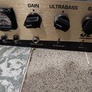 Detailní pohled na ovládací panel basového zesilovače zlaté barvy. Na panelu jsou černé ovratové knoflíky s popisky GAIN, ULTRABASS a BASS, dále je z…