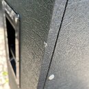 Detailní záběr na roh černého reproduktorového boxu s texturovaným povrchem. Na levé straně je vidět část černé plastové rukojeti připevněné šrouby.…