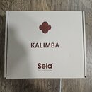 Fotografie znázorňuje uzavřenou kartonovou krabici v béžové barvě. Na horní straně je hnědý symbol čtyřlístku a nápisy KALIMBA a Sela® s doplňujícím…