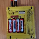 Žlutý plastový box s čtyřmi vloženými bateriemi značky Panasonic. Na horní části je štítek s nápisem MODEL: KO-1 a číslem 075055, vedle něj je nečite…