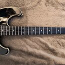 Černá elektrická kytara s tmavým hmatníkem a bílými vložkami ve tvaru křížků. Na těle je viditelný bílý pickguard a jeden snímač s kovovým krytem. Ná…