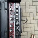 Detailní pohled na ovládací panel basového komba v červené barvě. Na panelu je nápis CUBE AD20DL BASS a logo COSM7 AMPLIFIER. Jsou zde různé ovladače…