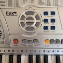 Detailní pohled na ovládací panel stříbrného elektronického klávesu s bílými a černými klávesami. V levé horní části je logo Fox instruments. Panel o…