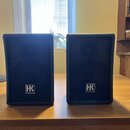 Dva černé kompaktní reproduktory s textilními maskami. Na spodní části obou reproduktorů je bílé logo ve čtverci s nápisem HK AUDIO. Povrchy jsou v d…