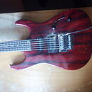 Elektrická kytara s červeným lakovaným tělem s viditnou strukturou dřeva. Nástroj má dva humbuckery, jeden ovládací knoflík a chromovaný tremolo syst…