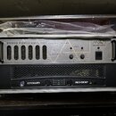 Stříbrný stereo výkonový zesilovač v hliníkovém transportním kufru. Horní část panelu obsahuje nápisy STEREO POWER AMPLIFIER NATURAL SOUND, logo PA s…