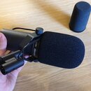 Černý dynamický mikrofon s nasazenou větrolamem z pěny, umístěný v černém držáku. Na těle mikrofonu je bílý nápis SHURE SM7B. Vedle mikrofonu leží sa…