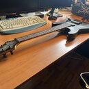 Černá elektrická kytara s matným povrchem a šesti strunami. Na hlavě hmatníku je zlatý nápis „Schecter“. Nástroj leží na dřevěném stole vedle počítač…