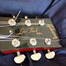 Detail hlavy elektrické kytary v černé barvě s červeným bokem. Na povrchu je zlatý nápis „Les Paul MODEL“ a vertikální logo „Gibson“. Jsou vidět stál…