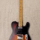 Elektrická kytara s tmelelovým povrchem v přechodu od černé přes červenou k žluté. Na hlavě hmatníku je nápis Fender TELECASTER. Kytara má černý pick…
