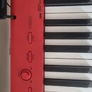Detailní pohled na červený digitální klavír s bílými a černými klávesami. Na ovládacím panelu jsou nápisy GRAND PIANO (STANDARD, MELLOW, BRIGHT), ELE…