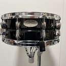 Černý malý buben s chromovaným obručováním a mechanismy, umístěný na stojanu. Na korpusu je oválný štítek s nápisy CONCERT, YAMAHA a JAPAN. Povrch ná…