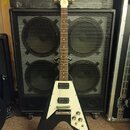 Černá elektrická kytara s bílým pickguardem a Flying V tvarem těla. Na hlavě je viditelné logo "Gibson". Na spodní části těla je nápis "Epiphone". Ky…