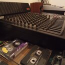 Černý mixovací pult s velkým množstvím potenciometrů a posuvníků. V horní části je umístěna naklonená panel s nápisem TASCAM. V popředí jsou v plasto…