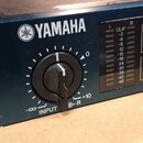 Detailní pohled na modrý kovový panel hudebního zařízení s bílým logem a nápisem YAMAHA. Na panelu je umístěn černý otočný knoflík s označením INPUT…