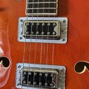 Detailní pohled na tělo oranžové polodubné elektrické kytary. Jsou vidět dva humbuckery s chromovanými kryty a bílými plastovými rámečky. V horní čás…