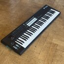 Černý MIDI kontrolér s klaviaturou, umístěný na dřevěné podlaze. Na horním panelu je bílé logo a nápis "novation launchkey". Nástroj obsahuje bílé a…