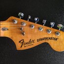 Fotografie zachycuje hlavu dřevěné elektrické kytary v přirozené barvě. Na hlavě je černý nápis Fender a STRATOCASTER®, pod ním menší text MADE IN US…