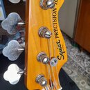 Fotografie zachycuje hlavu basové kytary z dřeva s žlutohnědým lakem. Na hlavě je černý nápis Squier PRECISION BASS® a pod ním menší nečitelný text.…