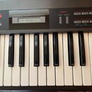 Detailní pohled na ovládací panel a klávesnici syntetizátoru v šedé barvě. Nad klávesami je nápis PROGRAMMABLE POLYPHONIC SYNTHESIZER JU-1 a displej…