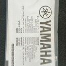 Fotografie znázorňuje bílý papírový manuál v průhledném plastovém obalu. Na titulní straně je svisle napsaný nápis YAMAHA a nad ním logo značky. Vedl…