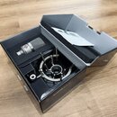 V černé krabici s pěnovou výplní je umístěn stříbrný kondenzátorový mikrofon s mřížkou a shock mount. Na těle mikrofonu je viditelné červené logo s b…