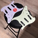 Dvě pickguardy do elektrické kytary v barvách světle fialové a krémové. Fialový pickguard má nalepenou červenou kulatou nálepku s nápisy Fender, FREE…
