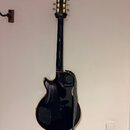 Černá elektrická kytara s vyvedeným okrajem v hnědé barvě, zavěšená na stěně. Na hlavě nástroje je viditelné logo Epiphone. Kytara má připojený šedý…