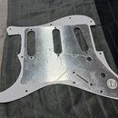 Bílý pickguard do elektrické kytary s hliníkovým stíněním na spodní straně. V pravé dolní části je viditelné logo "Genuine Fender". Povrch hliníkové…