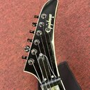Černá hlava elektrické kytary s bílým lemováním. Na povrchu je bílý nápis Epiphone. Jsou vidět šest ladicích kolíků a struny vedené k pražníku. Povrc…