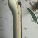 Detailní pohled na hranu krku bílé elektrické kytary. Na spoju hmatníku a krku jsou viditelné výrazné odloupení laku a praskliny. Povrch v této oblas…