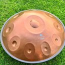 Kovový handpan v měděné barvě s několika vyrytými políčky a centrálním otvorem. Na povrchu je bílý nápis „Harmony Handpan“, pod ním logo ve tvaru kyt…